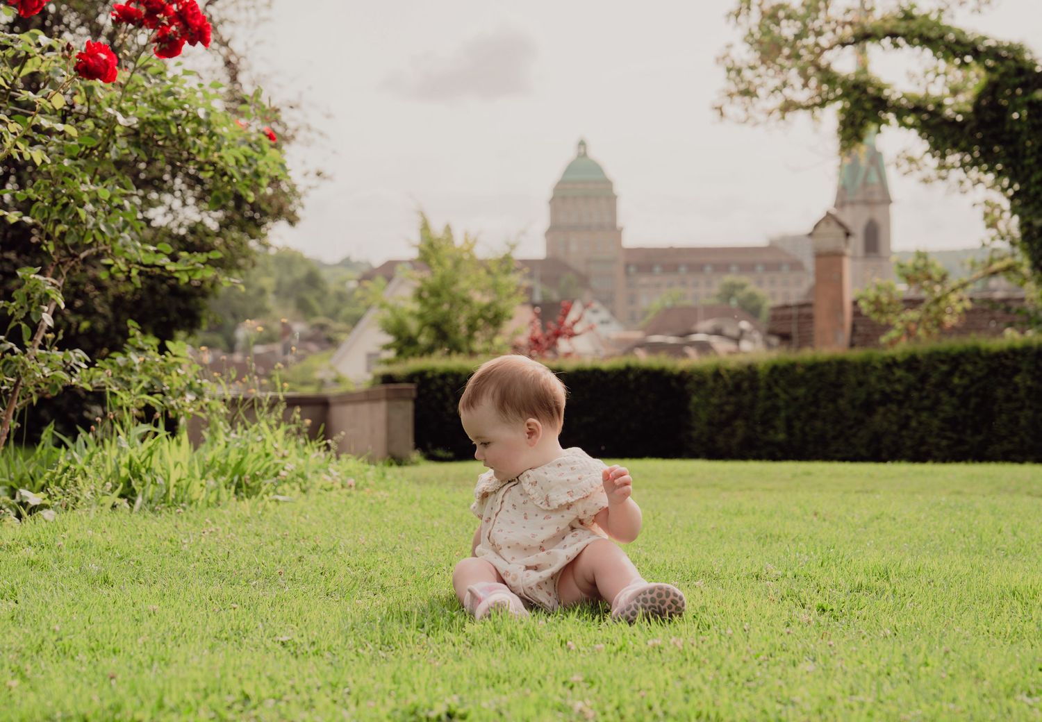Baby in cream floral romper sitting on green grass with historic domed building backdrop.