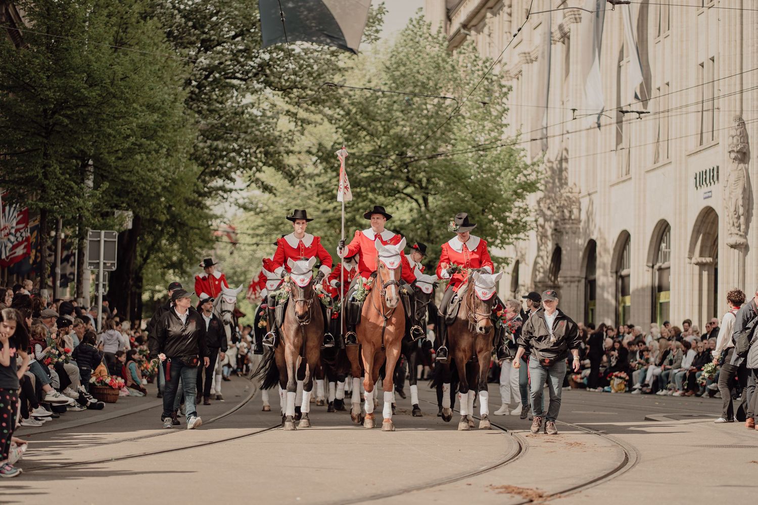 Riders in red uniforms on horseback lead a traditional parade through a historic European city street.