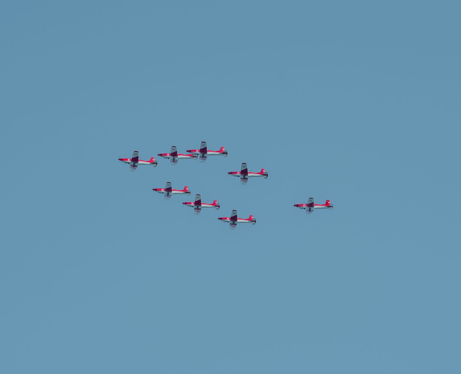 Nine red and white aerobatic planes flying in formation against clear blue sky.