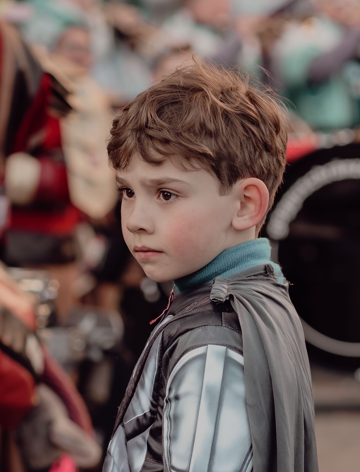 A young boy in a silver armored costume with a cape watches a festive parade.
