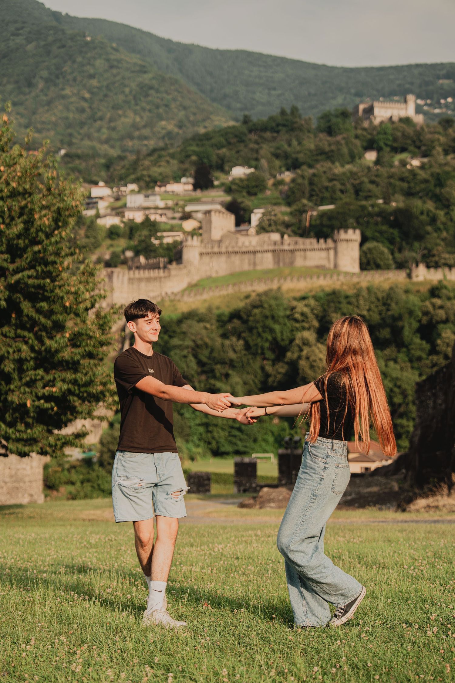 Couple joyfully spinning and holding hands on a green hillside with a medieval castle backdrop.