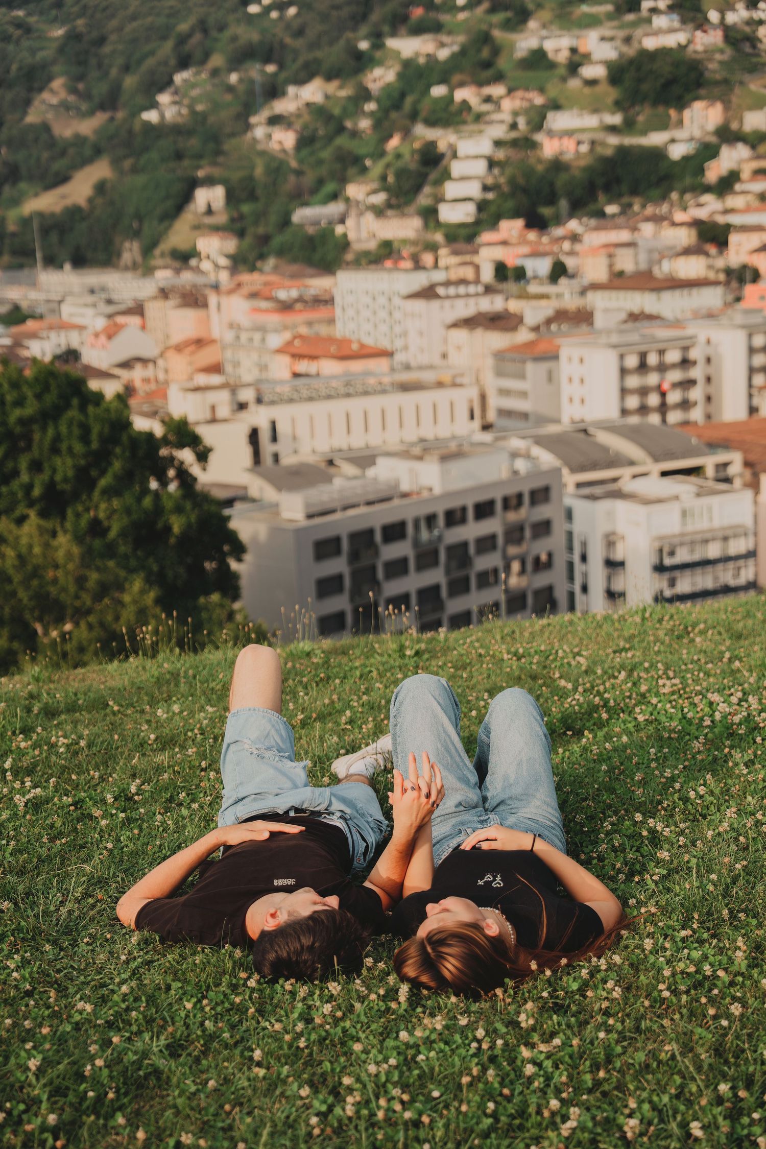A romantic couple lying on a flower-dotted hillside holding hands with a picturesque town visible below.