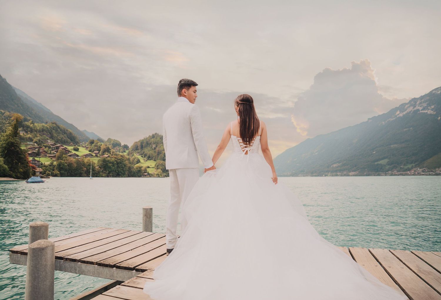 Newlywed couple stands hand in hand on lakeside dock admiring scenic alpine mountains and turquoise water.