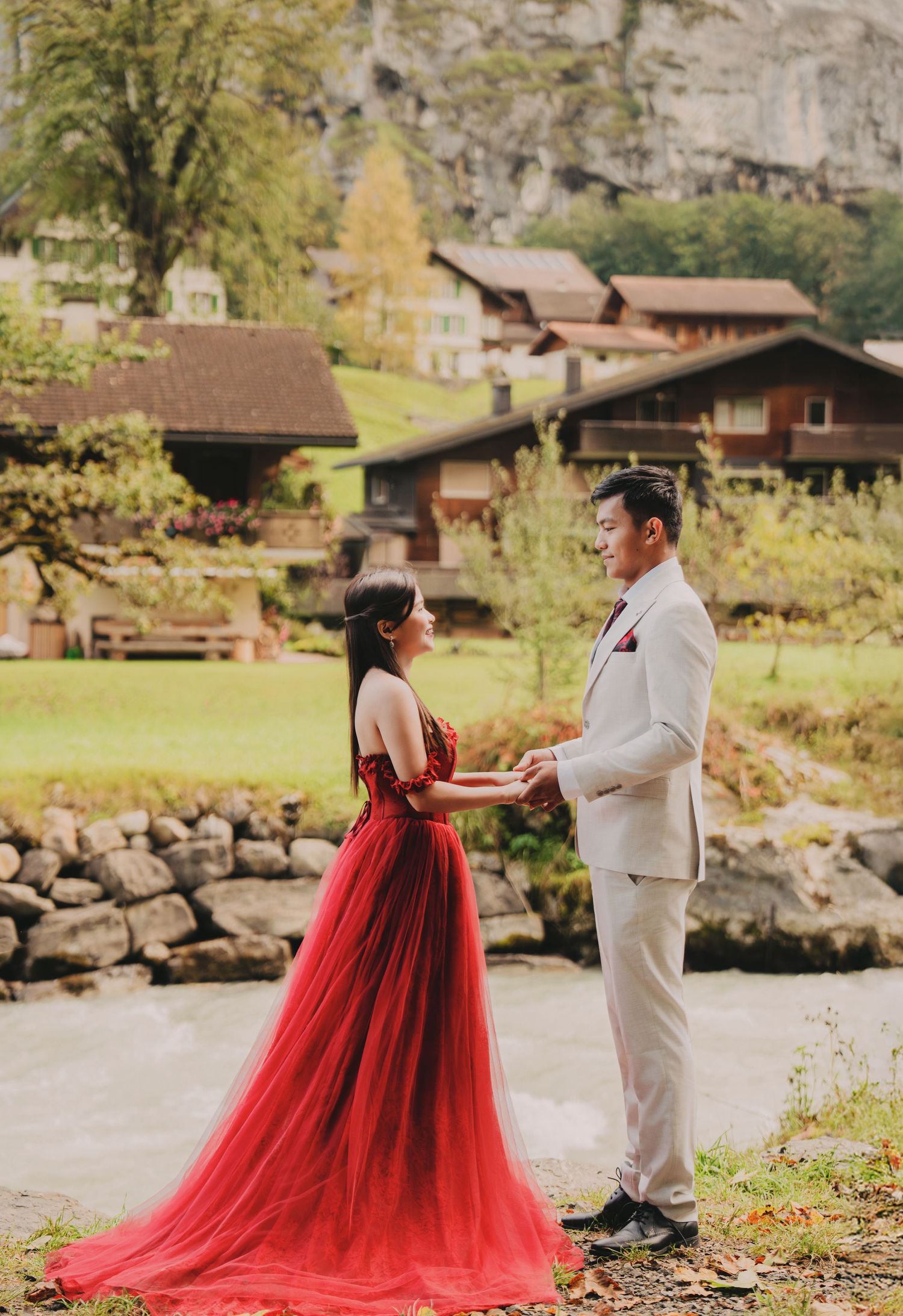 Couple exchanging vows holding hands in red tulle gown beside stone wall in Swiss village landscape.