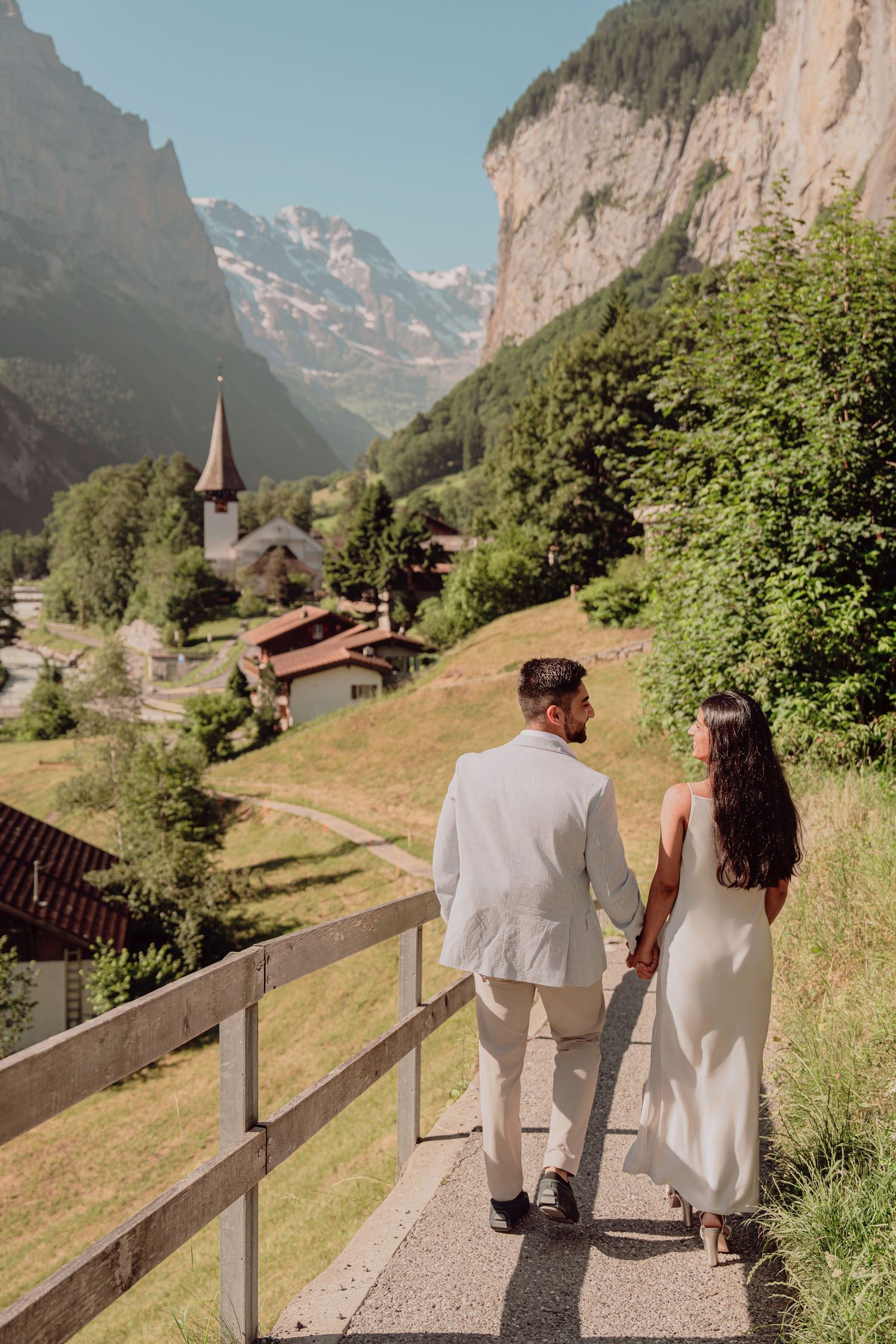Couple walking hand in hand on a path with Lauterbrunnen valley and church below.
