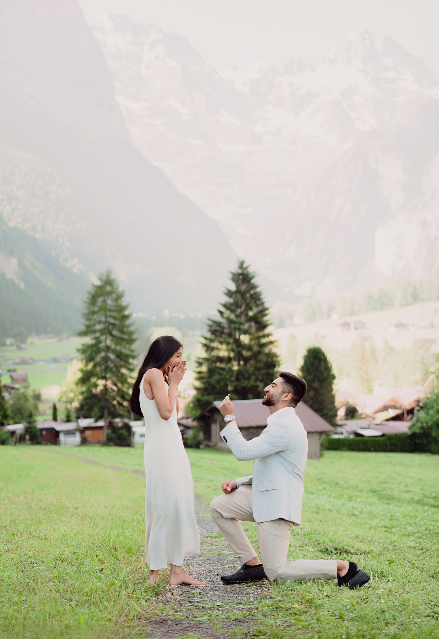 Man proposing on one knee to surprised woman in scenic Swiss alpine valley with mountains behind.