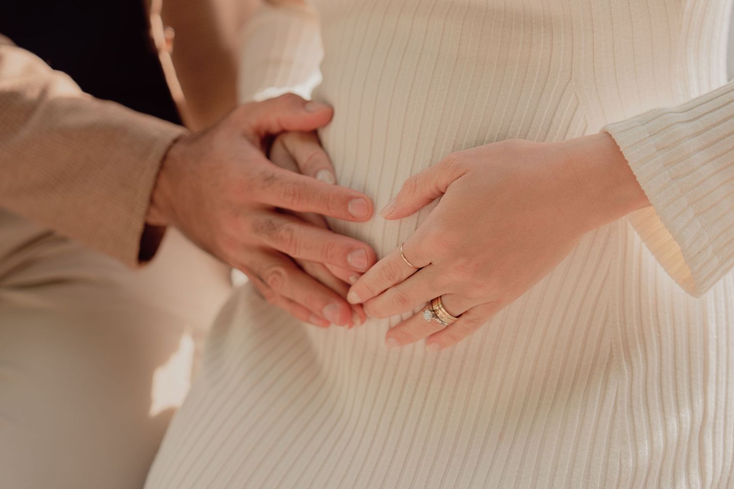 Couple tenderly holds pregnant belly together, woman wearing gold ring, both in neutral tones.