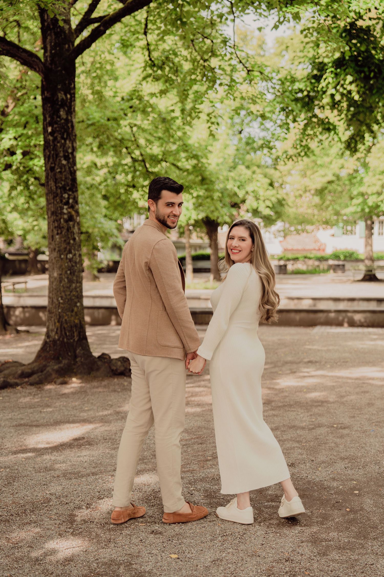 Smiling pregnant couple holding hands and posing in a sunny park, woman in cream dress, man in beige blazer.
