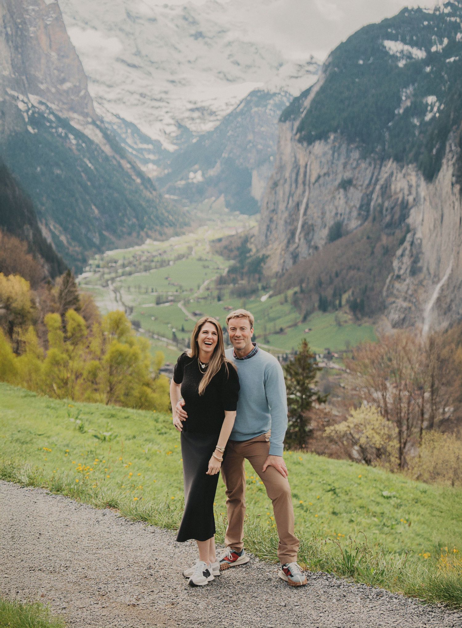 Smiling couple holds hands on mountain path with breathtaking Swiss Alps valley view behind them.