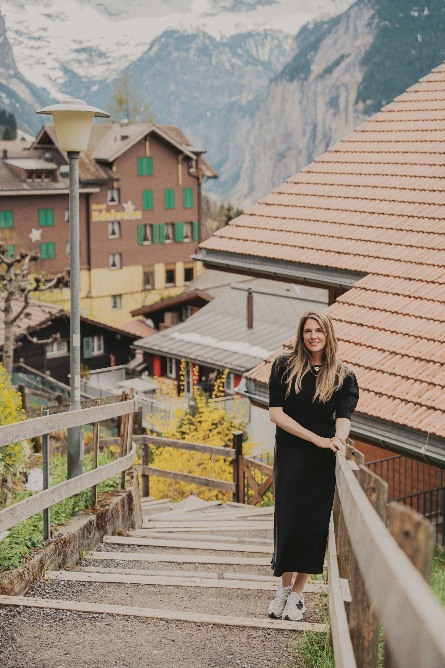 Woman in black dress standing on wooden staircase in a Swiss Alpine village with mountain views.