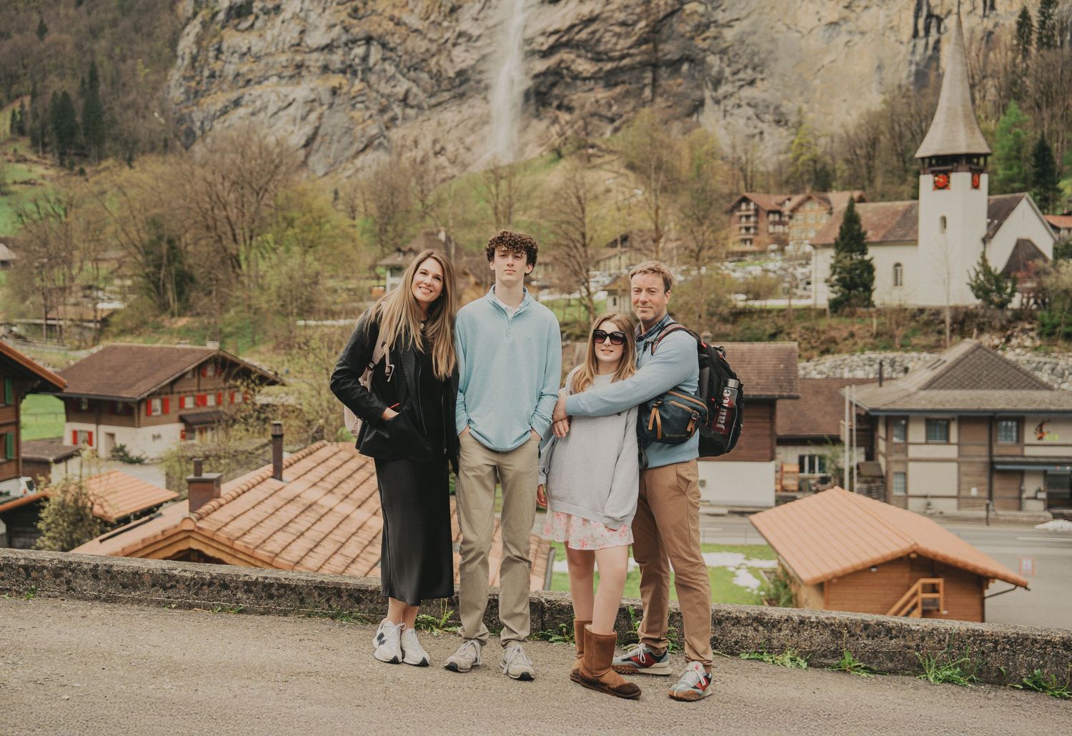 Family of four posing with Lauterbrunnen waterfall and Swiss village church visible in the background.