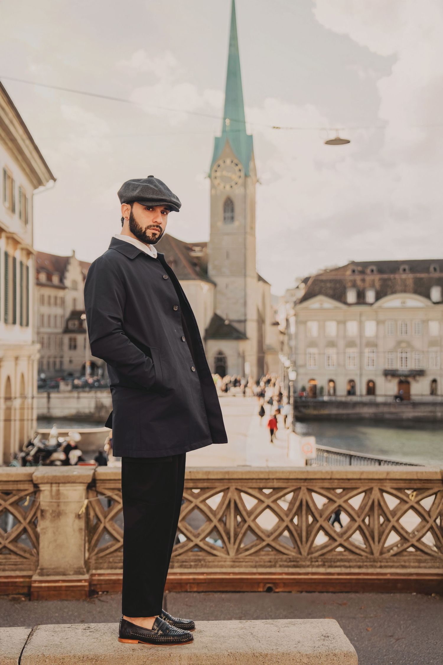 Fashionable bearded man in dark trench coat and flat cap stands on Zurich bridge overlooking the old town.