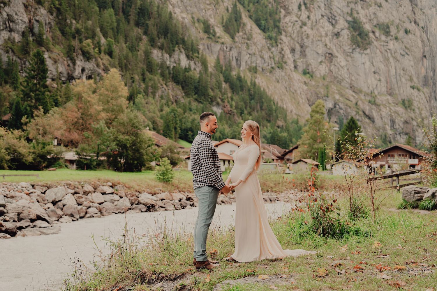 Pregnant couple holding hands facing each other in romantic maternity shoot with alpine mountains and chalets behind.