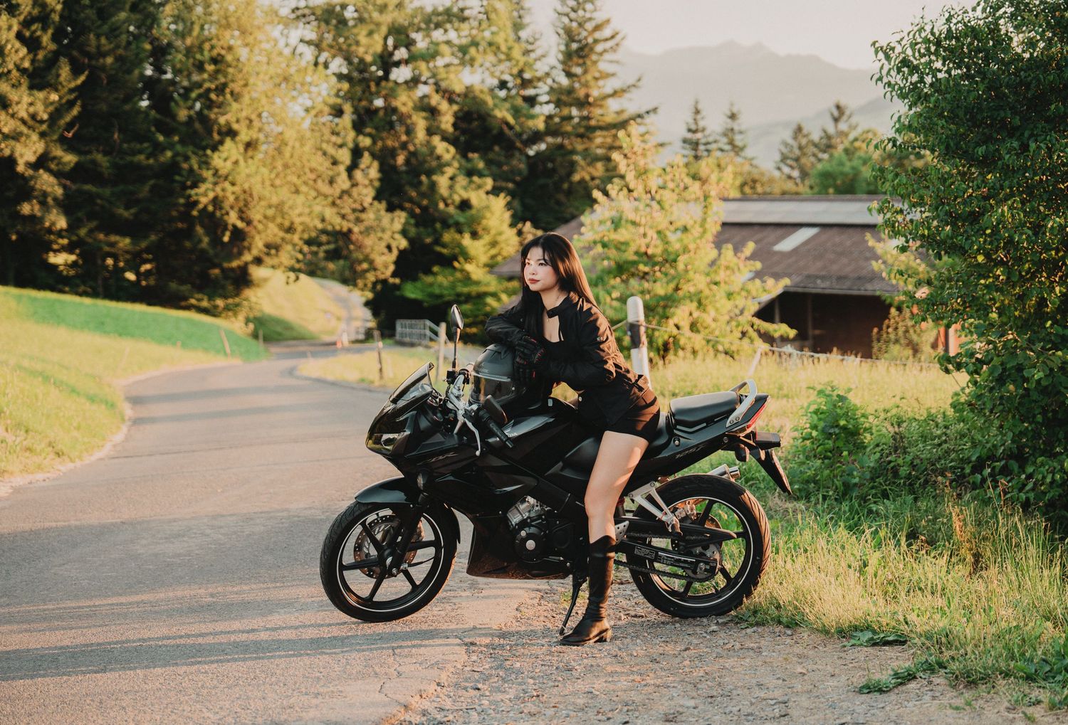 Young woman in black outfit and knee-high boots posing on a black Honda motorcycle on a countryside road.