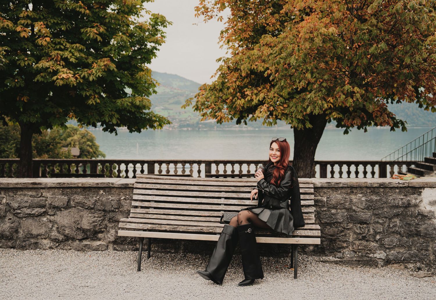 Woman in black outfit sitting alone on a park bench with lake and autumn trees in background.