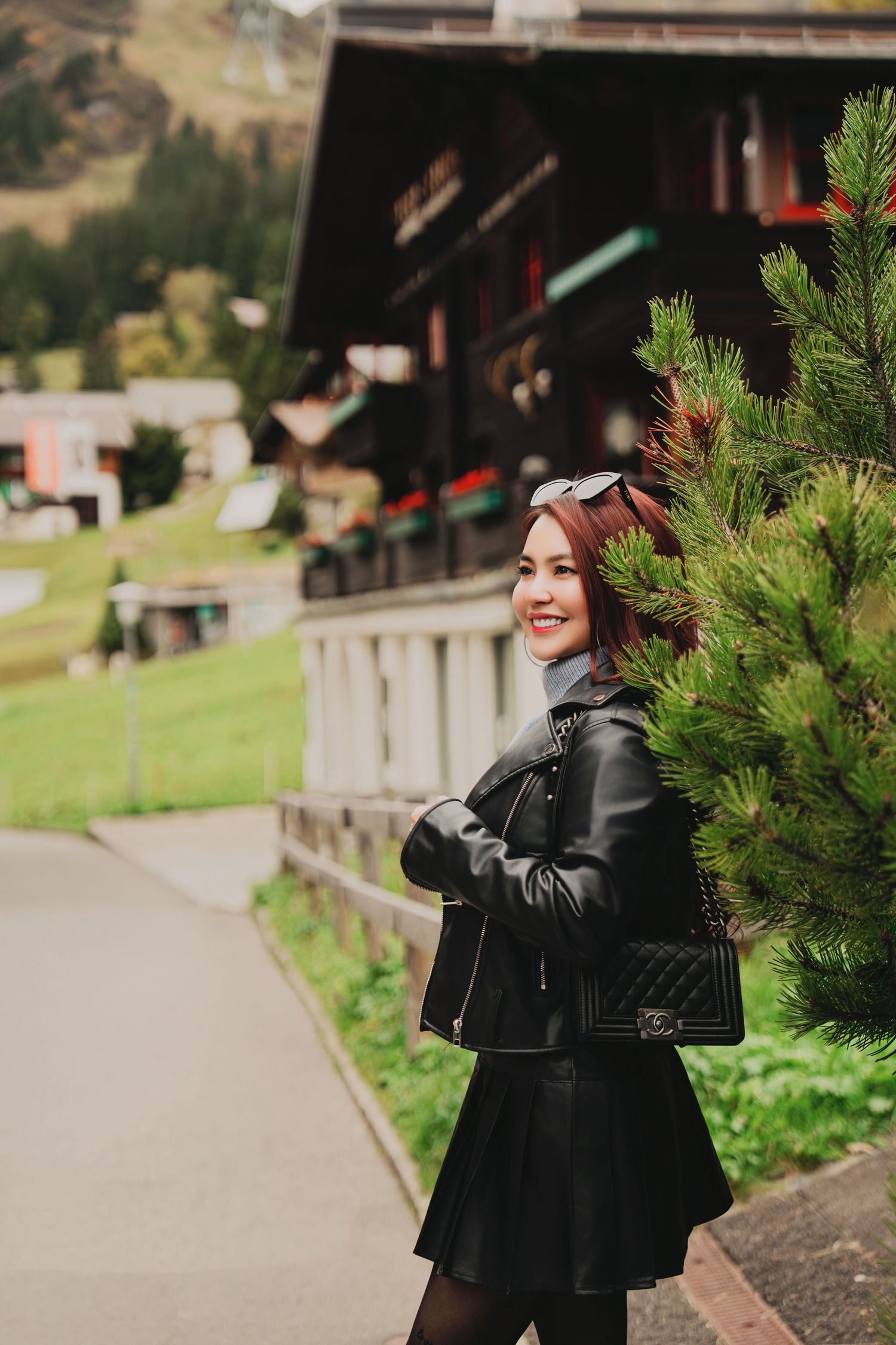 Smiling woman in black leather jacket and skirt poses beside an evergreen tree in a Swiss mountain village.