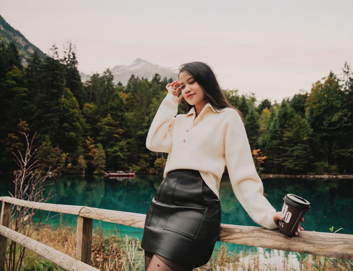 Woman in cream fuzzy sweater and black leather skirt holding coffee near wooden fence at alpine lake.