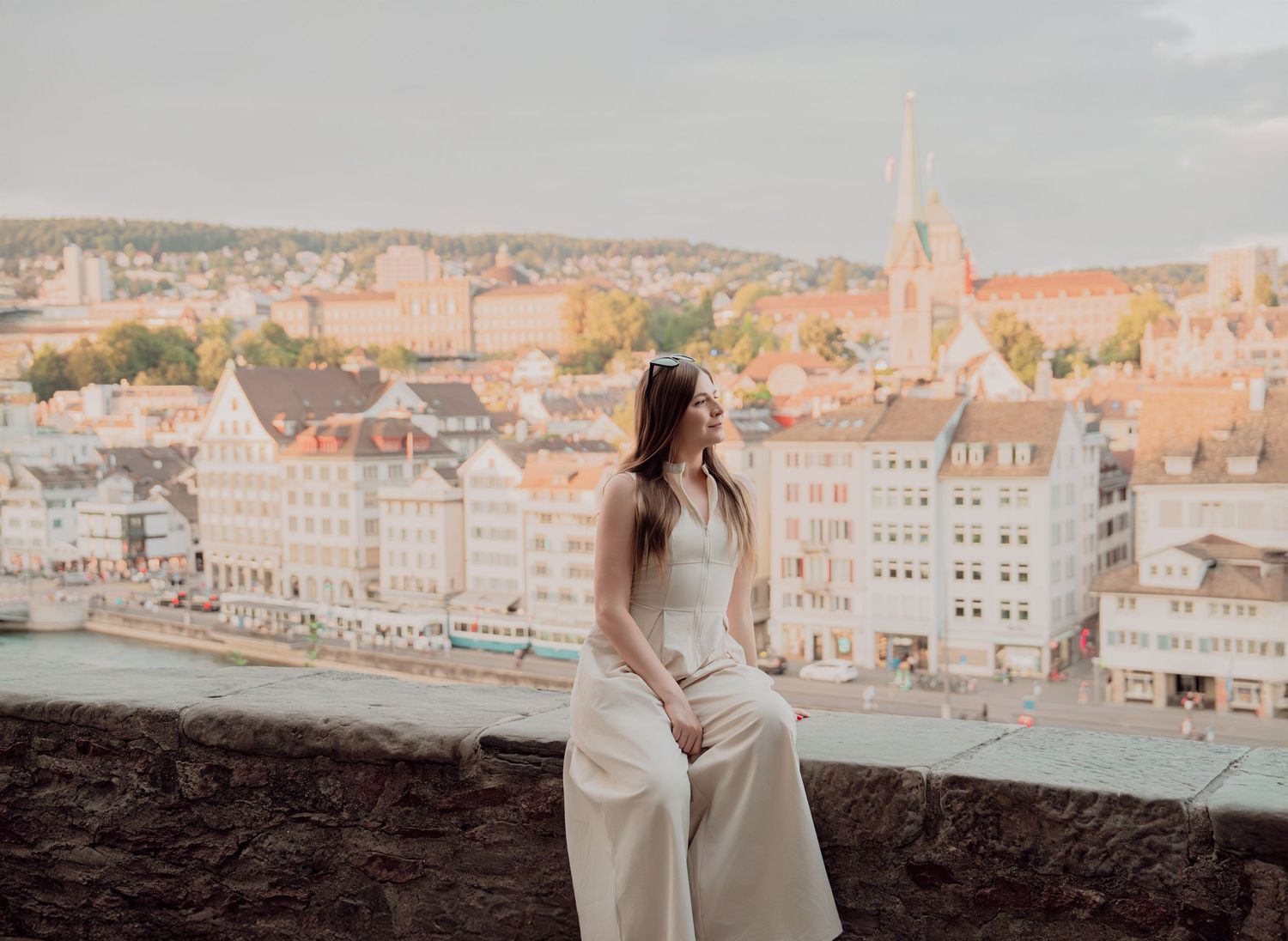 Young woman in cream outfit posed on stone wall overlooking colorful European city with cathedral spire.