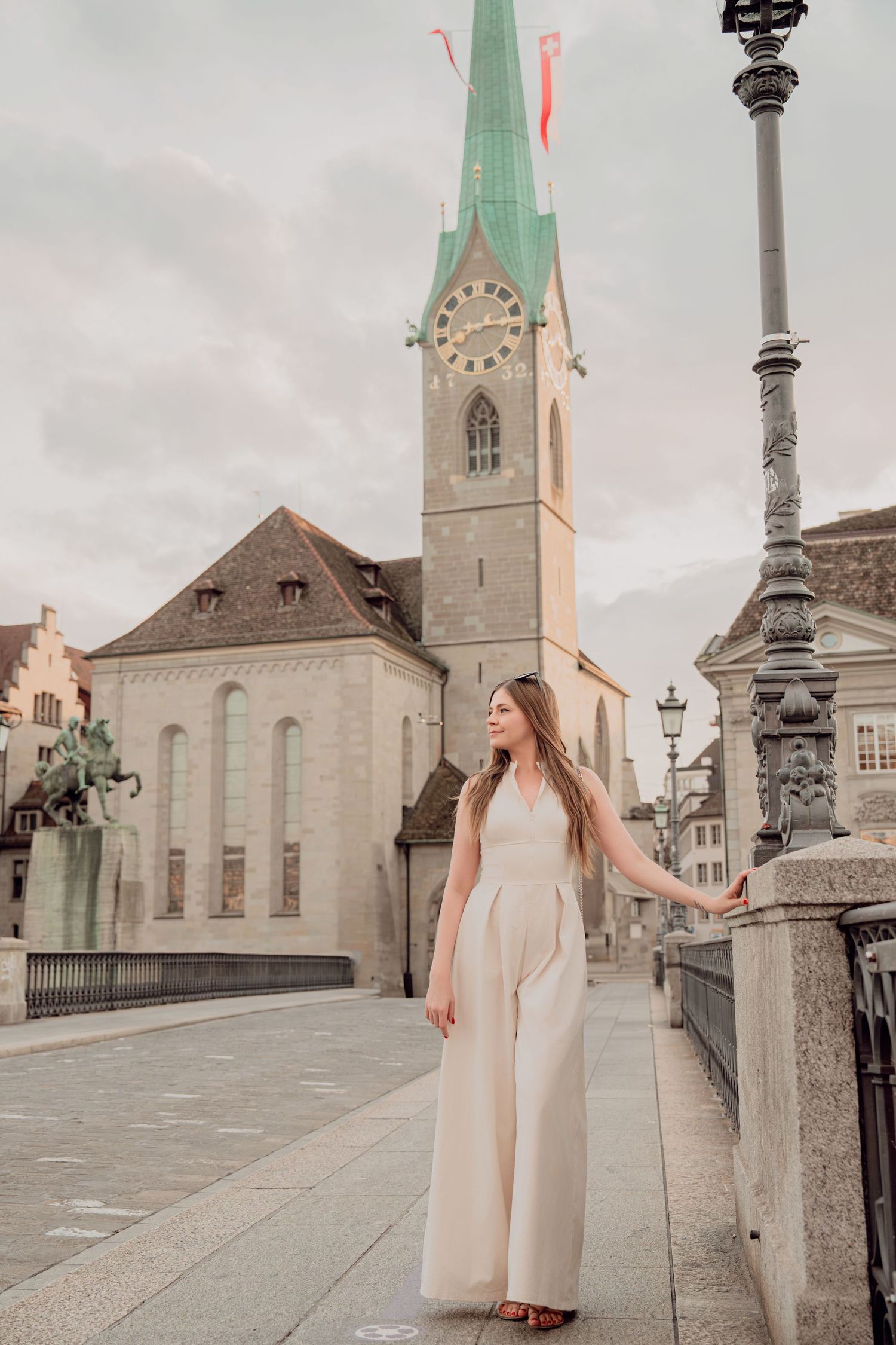 Woman in cream jumpsuit poses by ornate lamppost with Fraumunster Church clock tower in Zurich, Switzerland.
