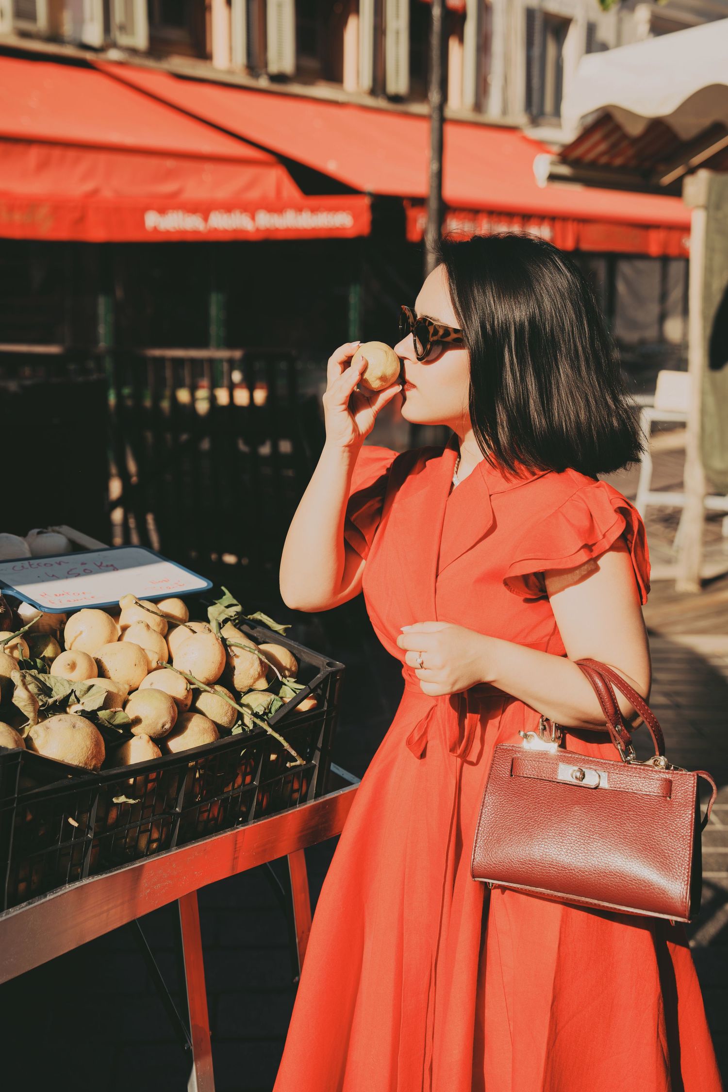 Chic woman in red dress and sunglasses smelling fresh fruit at outdoor market stall.