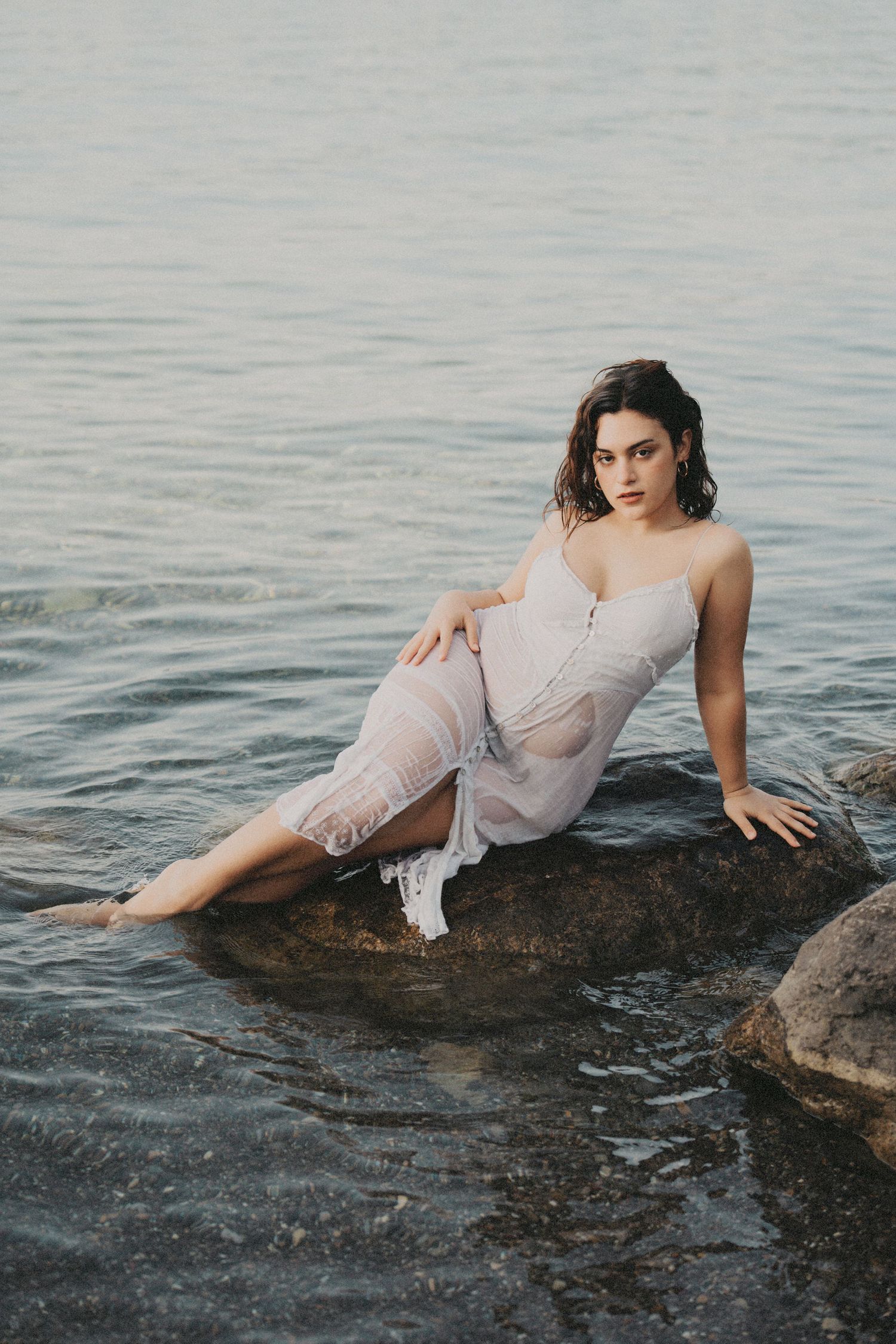 Woman in sheer white dress sitting on rocks surrounded by calm water.