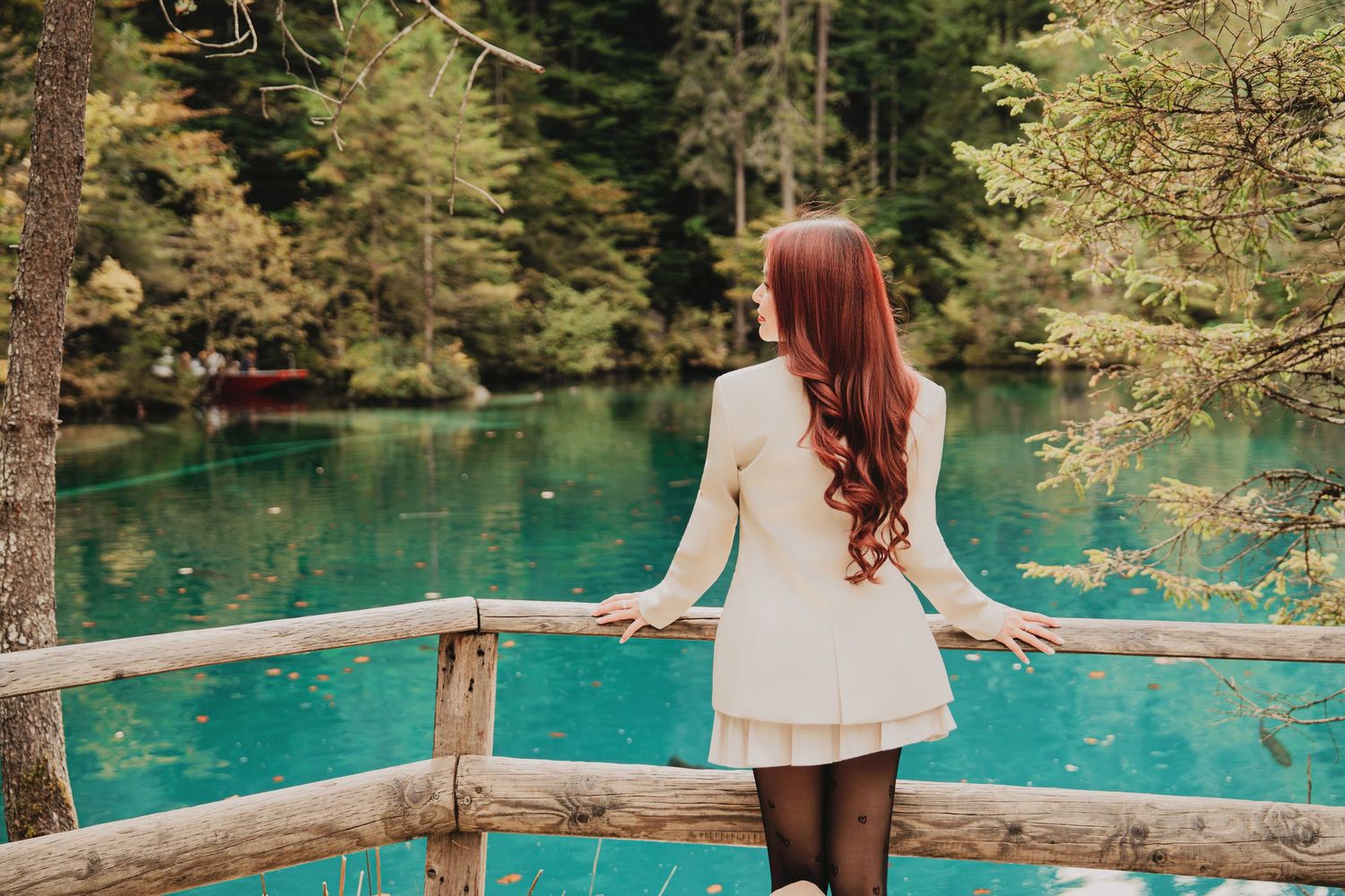 Red-haired woman in white dress gazing at turquoise alpine lake from wooden fence.