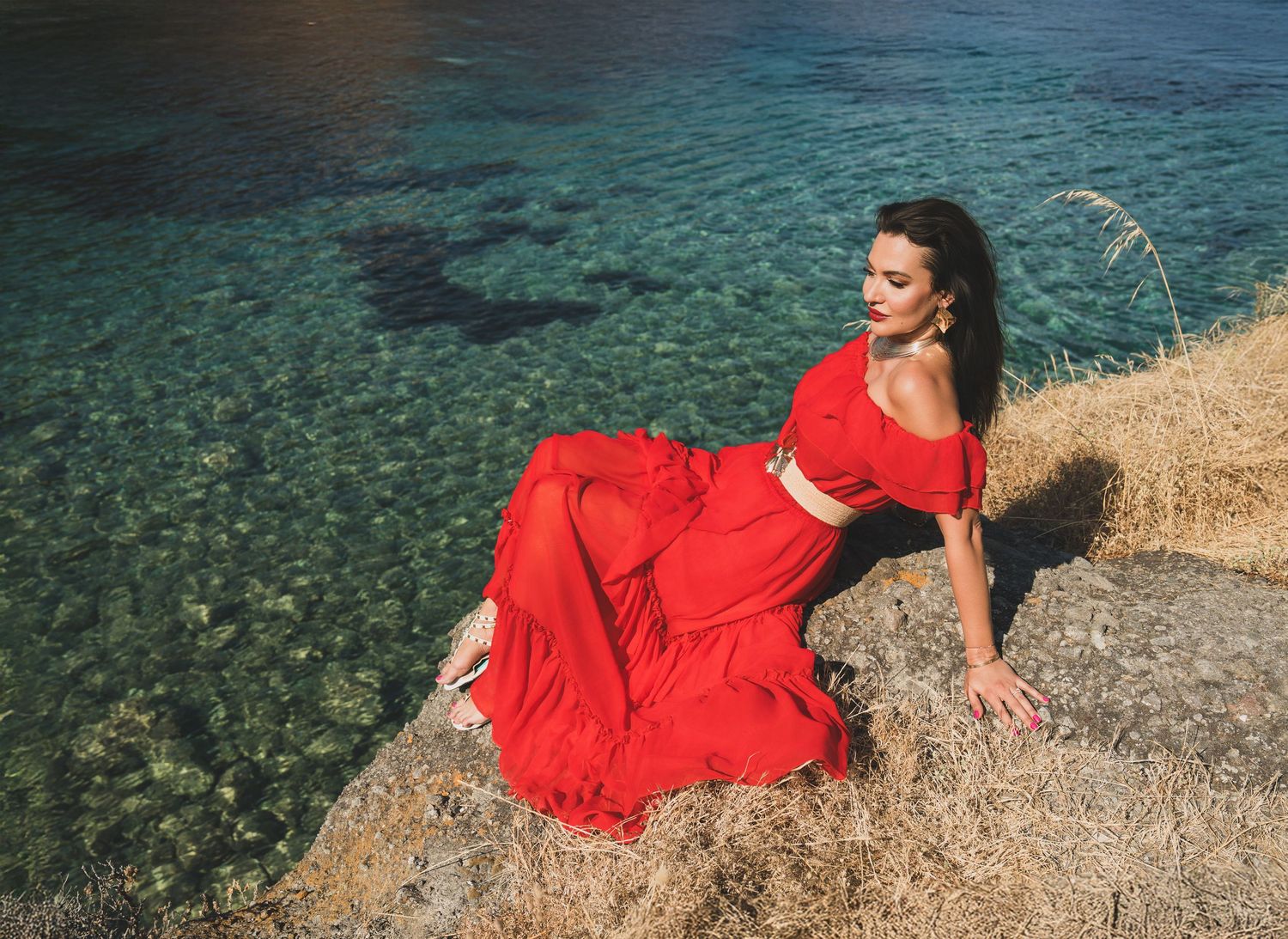 Woman in red crop top and maxi skirt reclining on coastal rocks above crystal clear water.