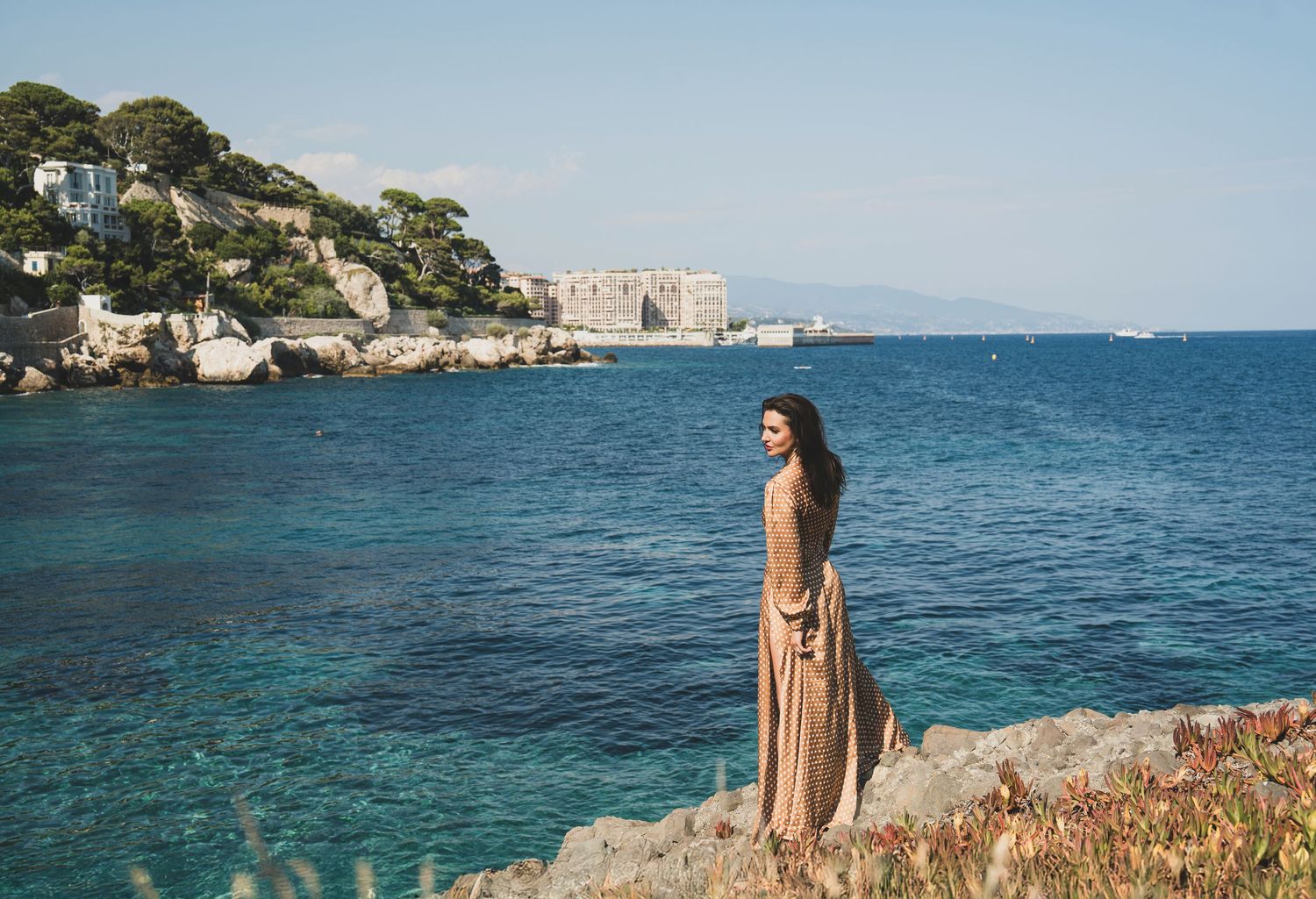 Woman in elegant beige maxi dress standing on rocky Mediterranean coastline with turquoise sea views.