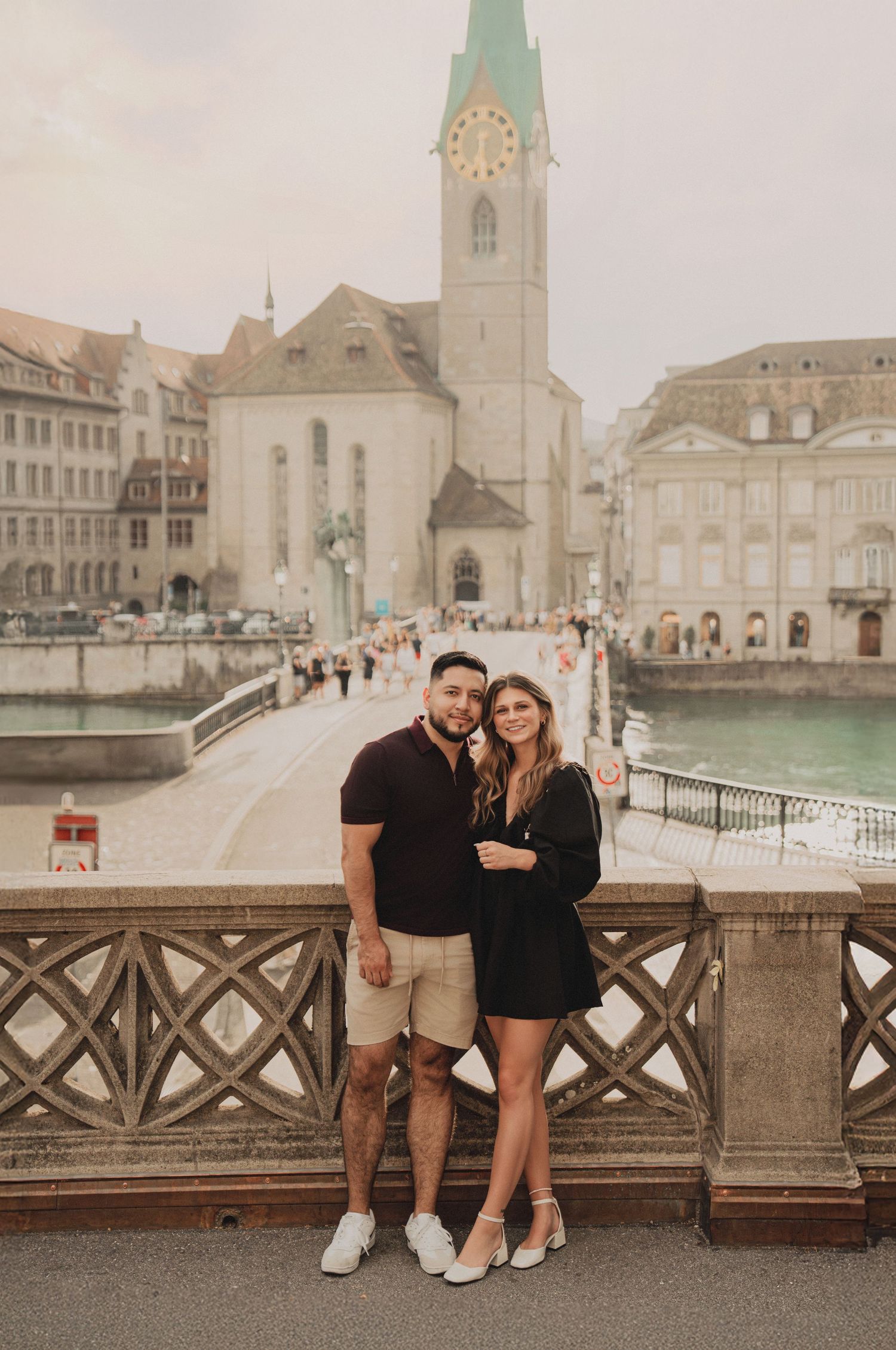 Couple embracing on Munsterbrucke bridge in Zurich with Fraumunster church and river in background.