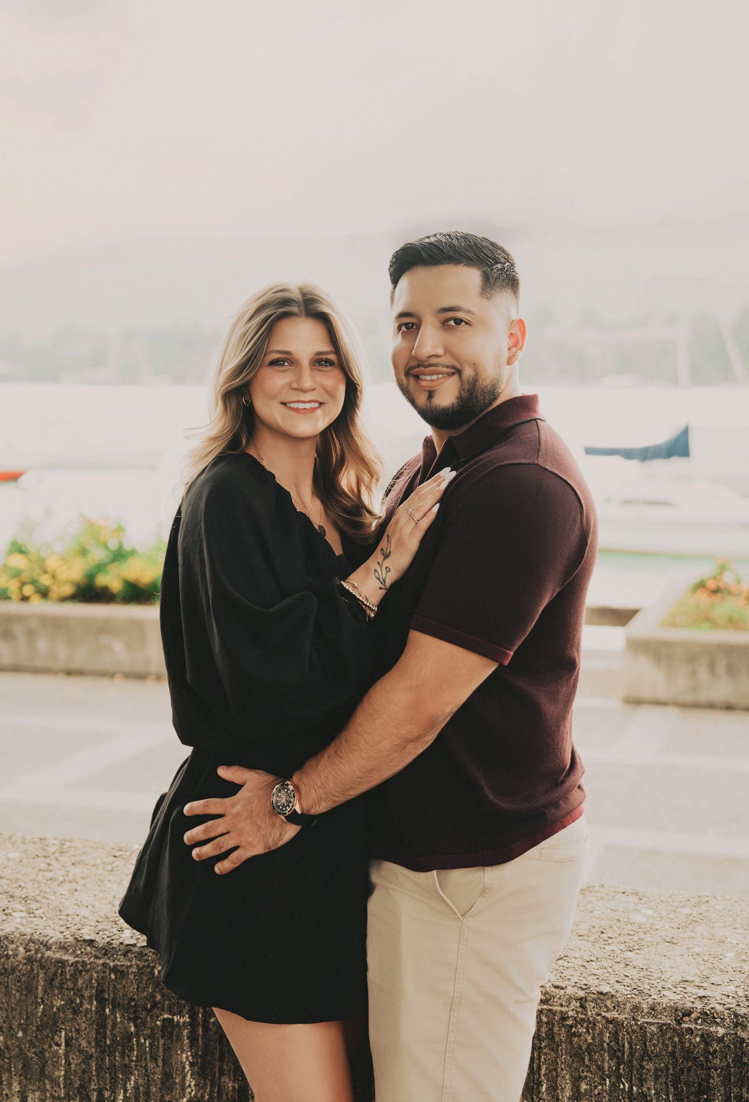 Couple embracing near a lakeside wall in Zurich, woman in black dress, man in burgundy polo shirt.