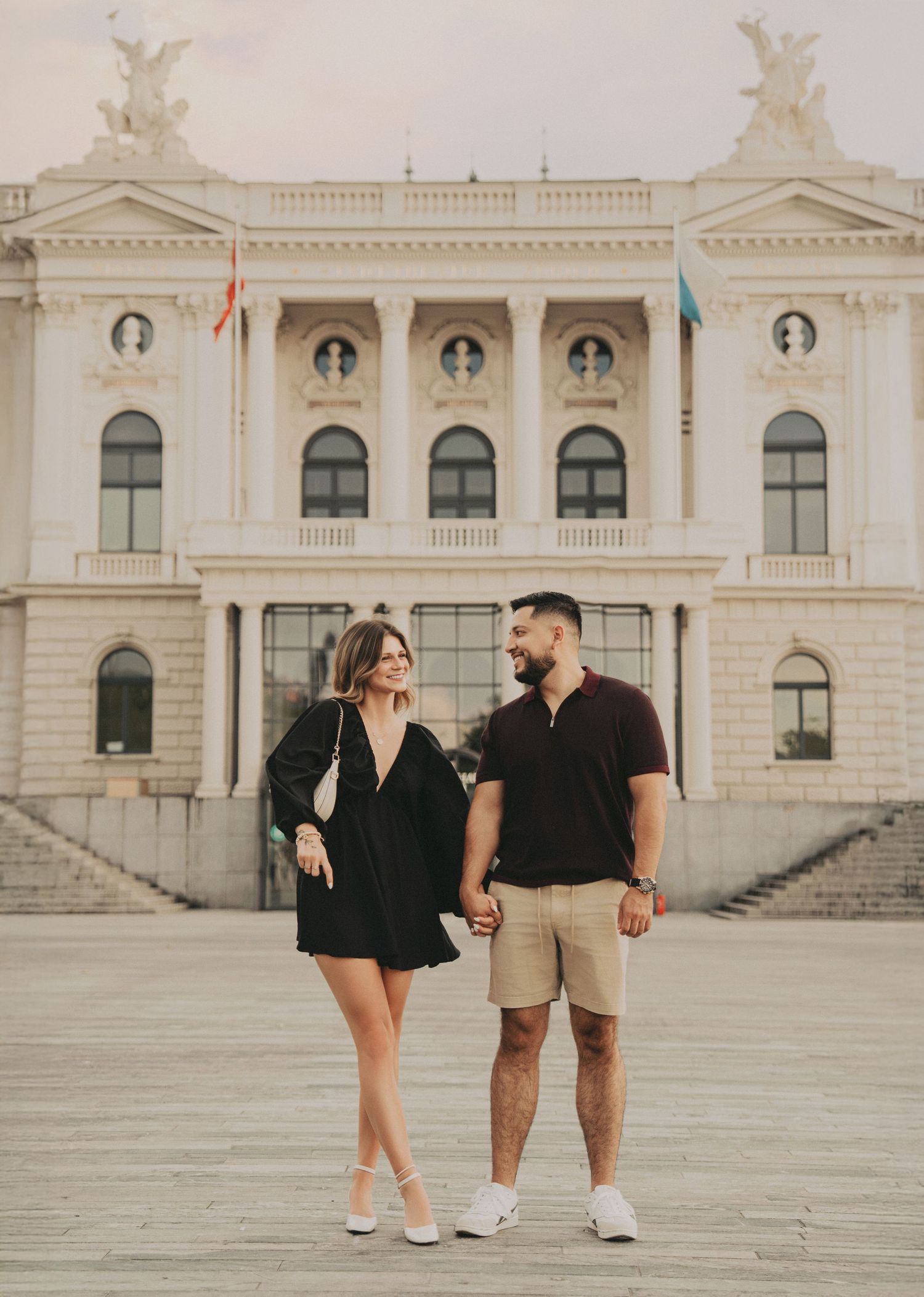 Couple holding hands and gazing at each other before a grand classical European building.