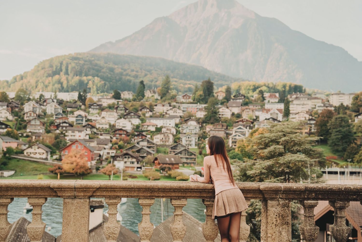 Young woman leaning on ornate stone balustrade admiring scenic Alpine village and mountain view.