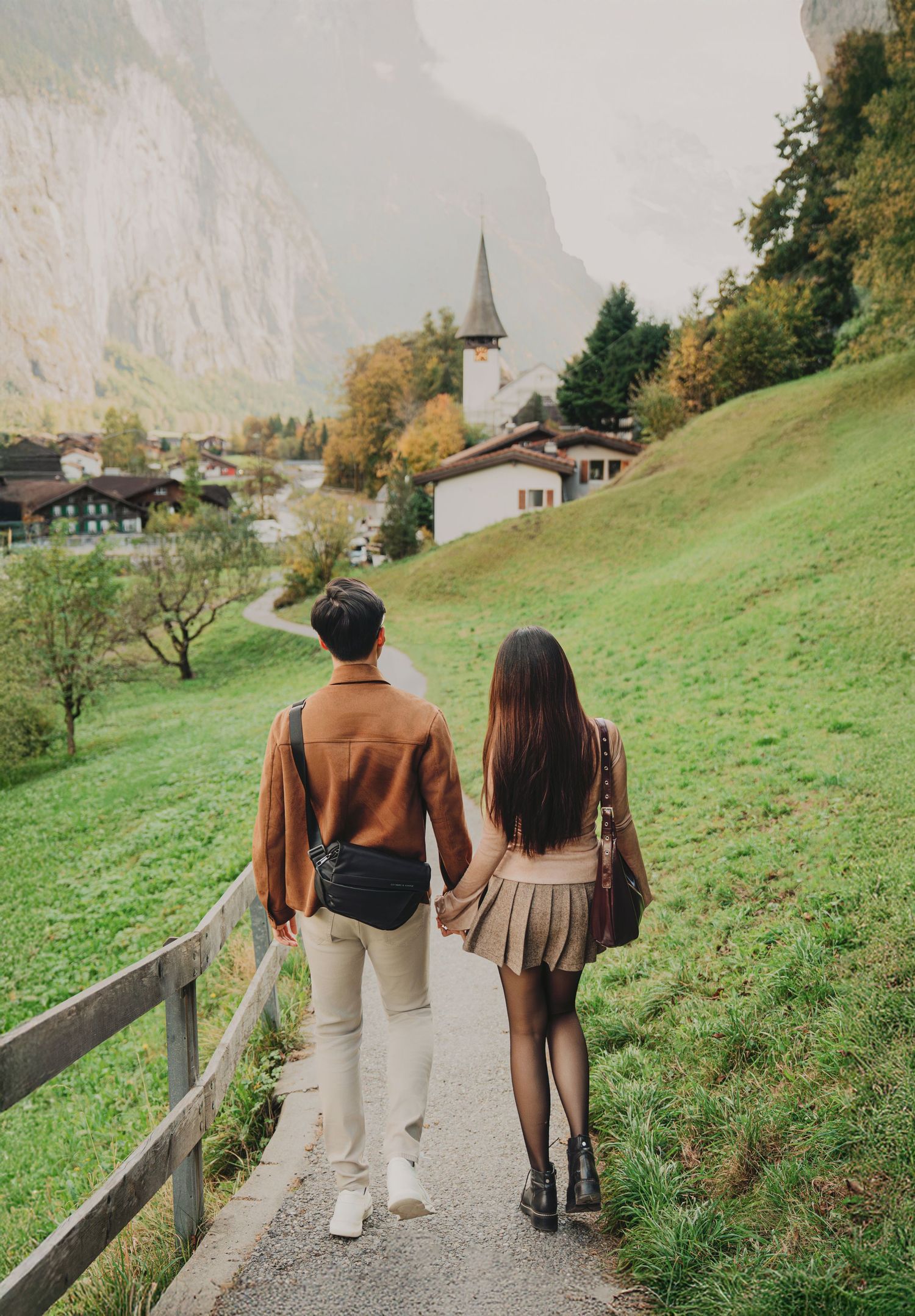 Couple holding hands walking on a path through a lush Swiss alpine village with a church steeple.