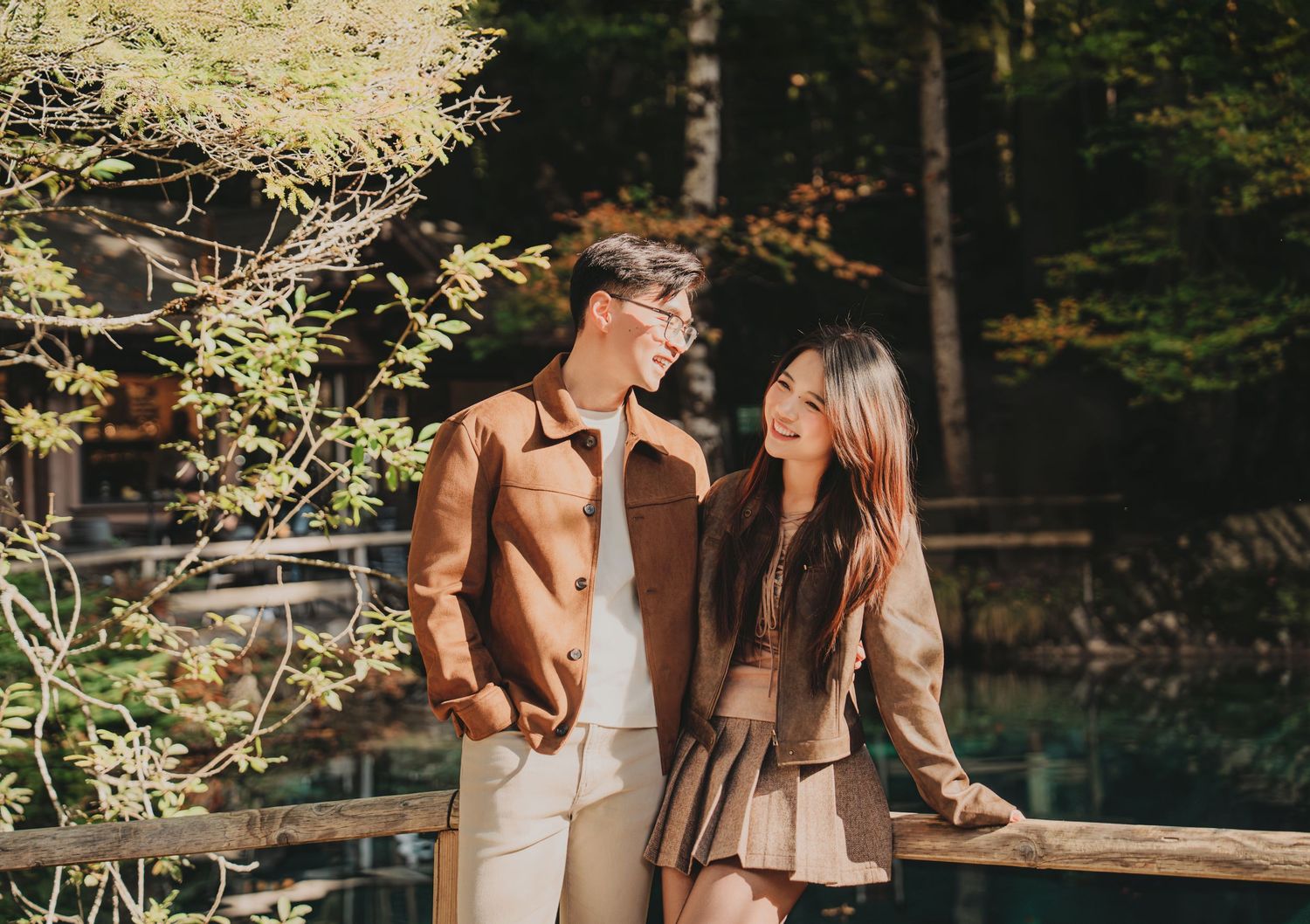 Smiling couple holding hands on a wooden bridge surrounded by autumn forest trees.
