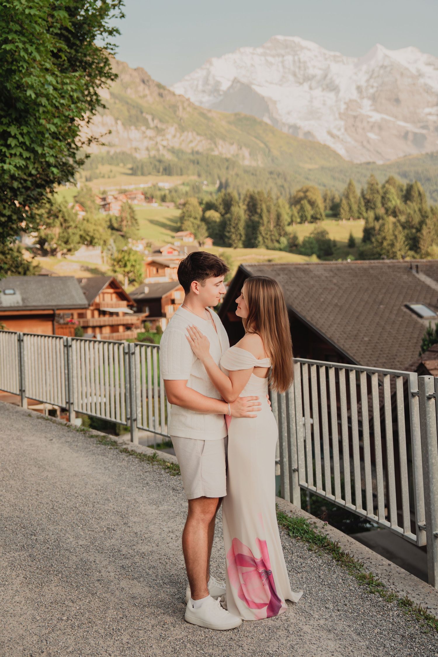 Couple in matching neutral outfits embracing on a mountain road with beautiful Swiss Alps village scenery behind them.