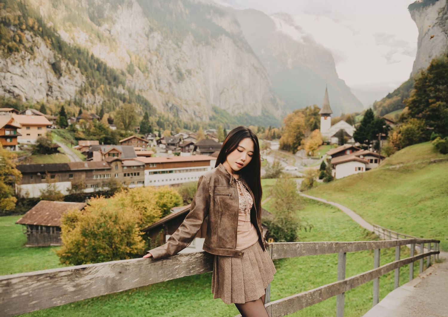 Young woman in brown jacket leans on wooden fence with colorful Swiss mountain village in background.