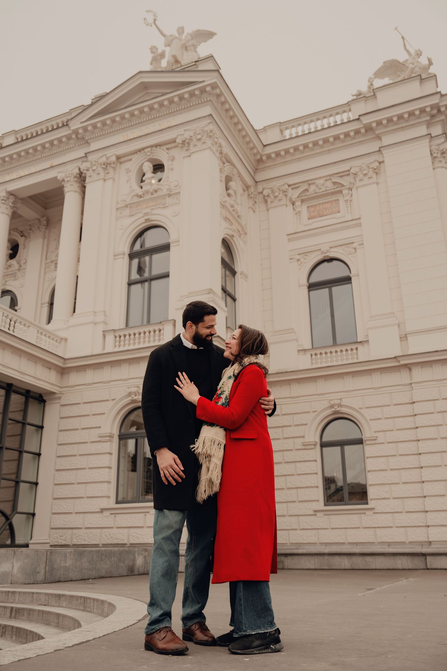 Couple shares a romantic moment in front of a grand white neoclassical building, woman in red coat.