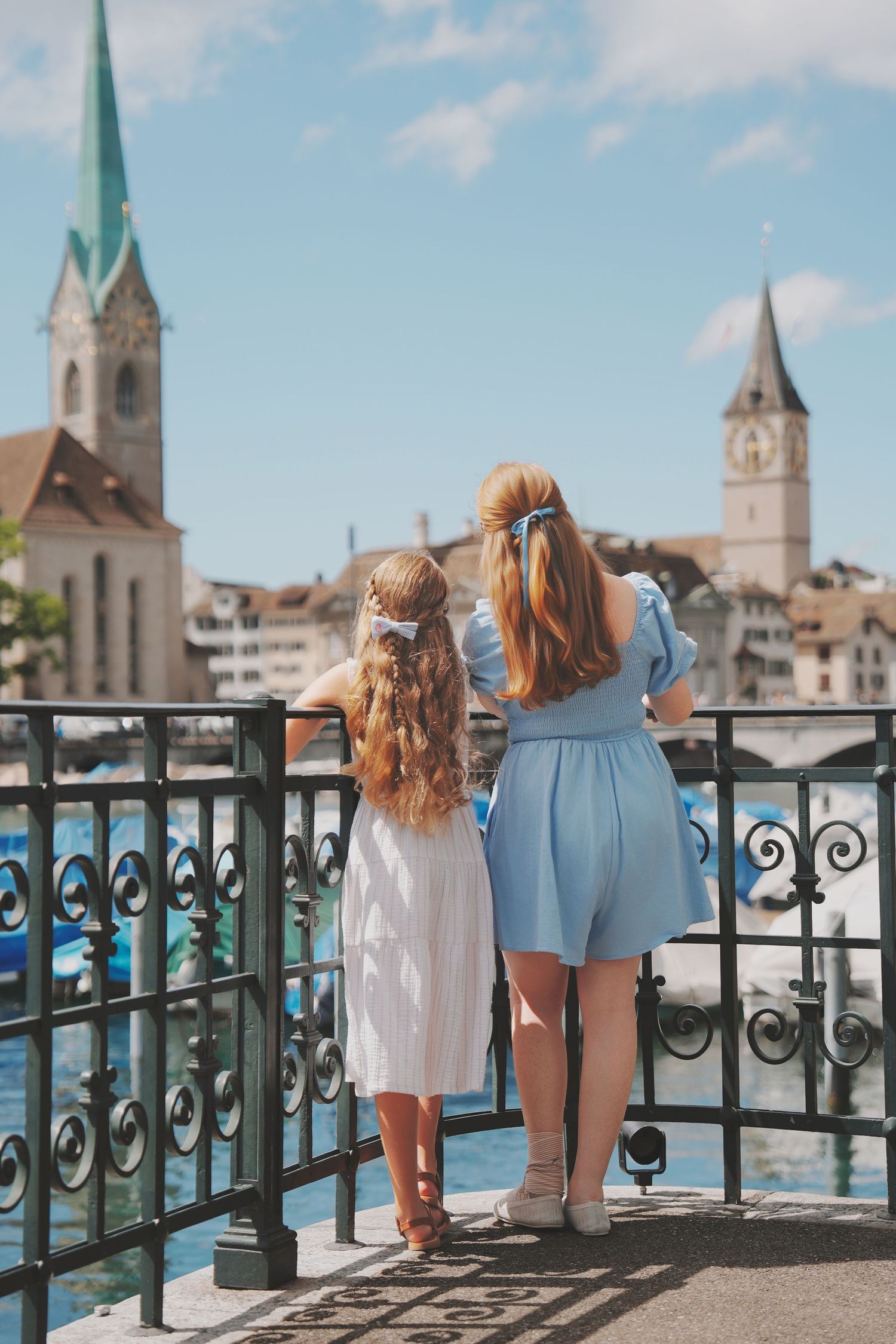 A mother and daughter in matching blue and white dresses admire the Zurich skyline from a riverside bridge.