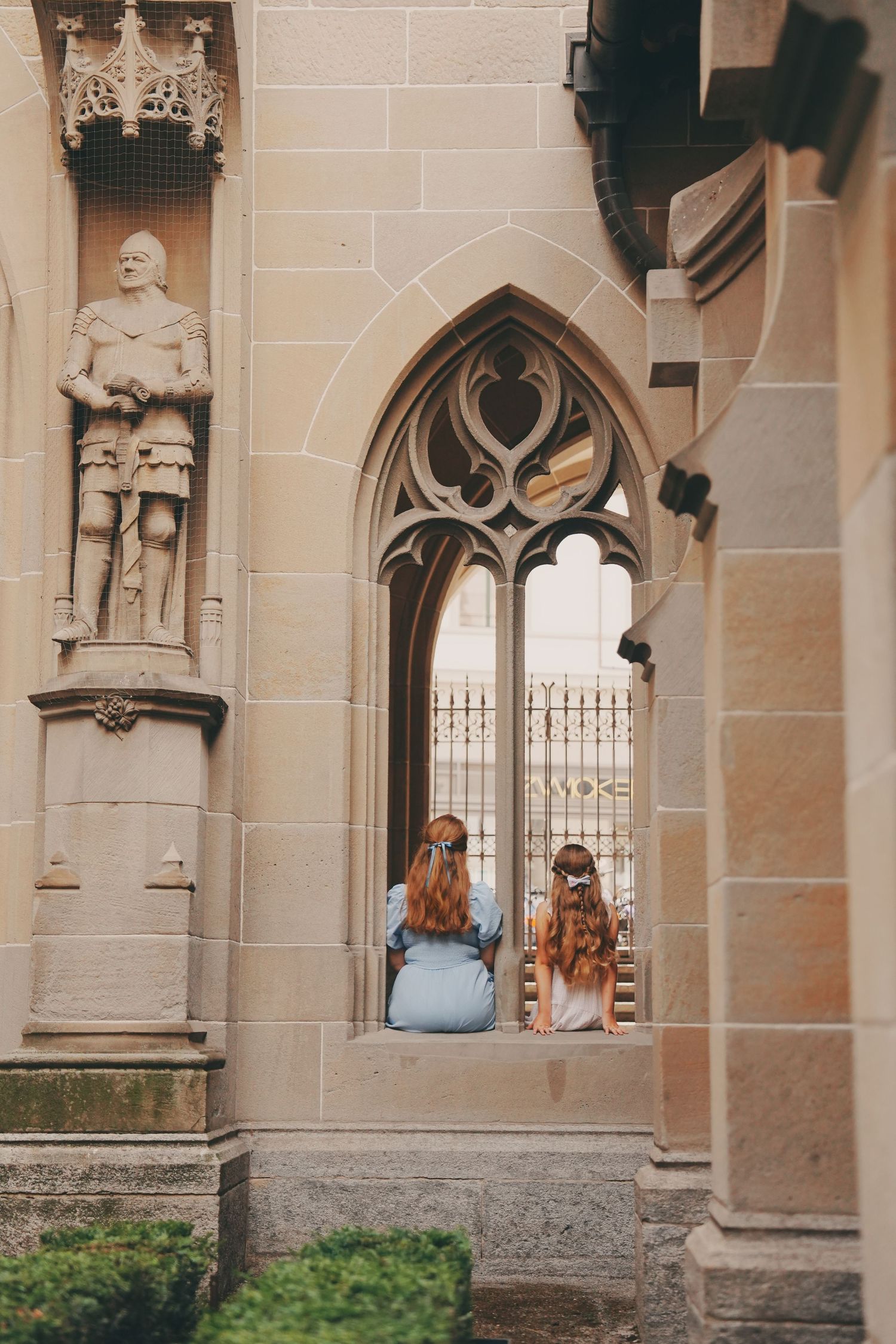 Mother and daughter sitting in Gothic arched window of historic stone church with knight statue.