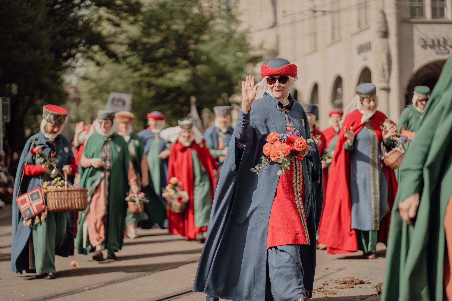 Parade participant in blue robe waves while carrying orange roses at a traditional street festival.