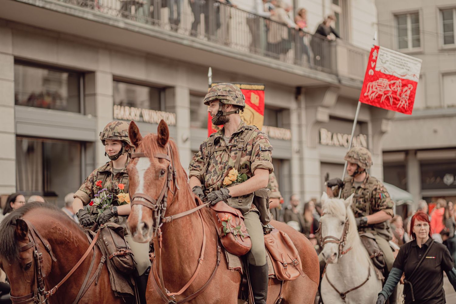 Three military equestrians carrying red flags ride through a festive street parade.