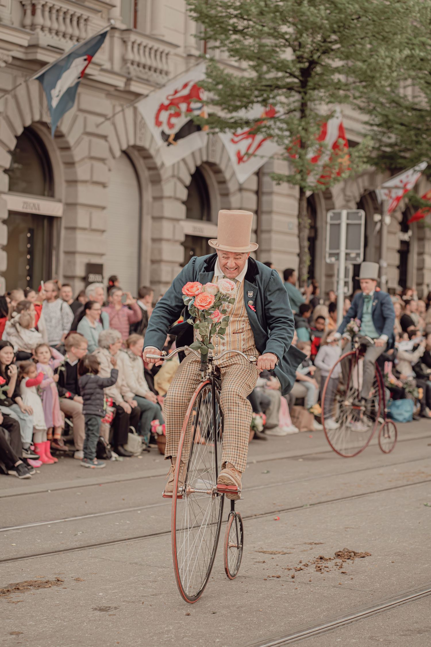 Man in vintage attire rides a penny-farthing bicycle holding roses at a street parade.