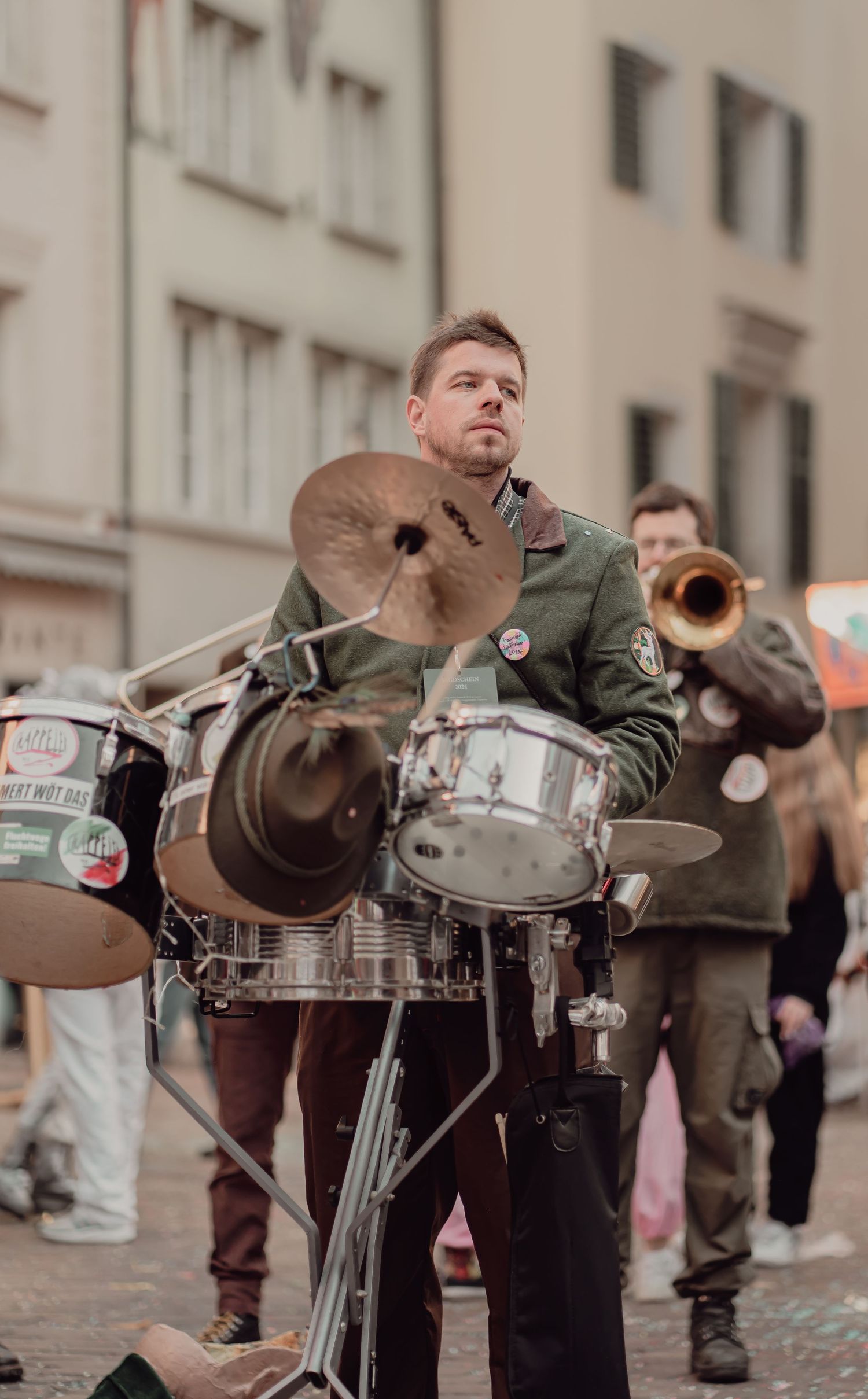 Street musician playing drums at outdoor carnival with trumpet player behind.