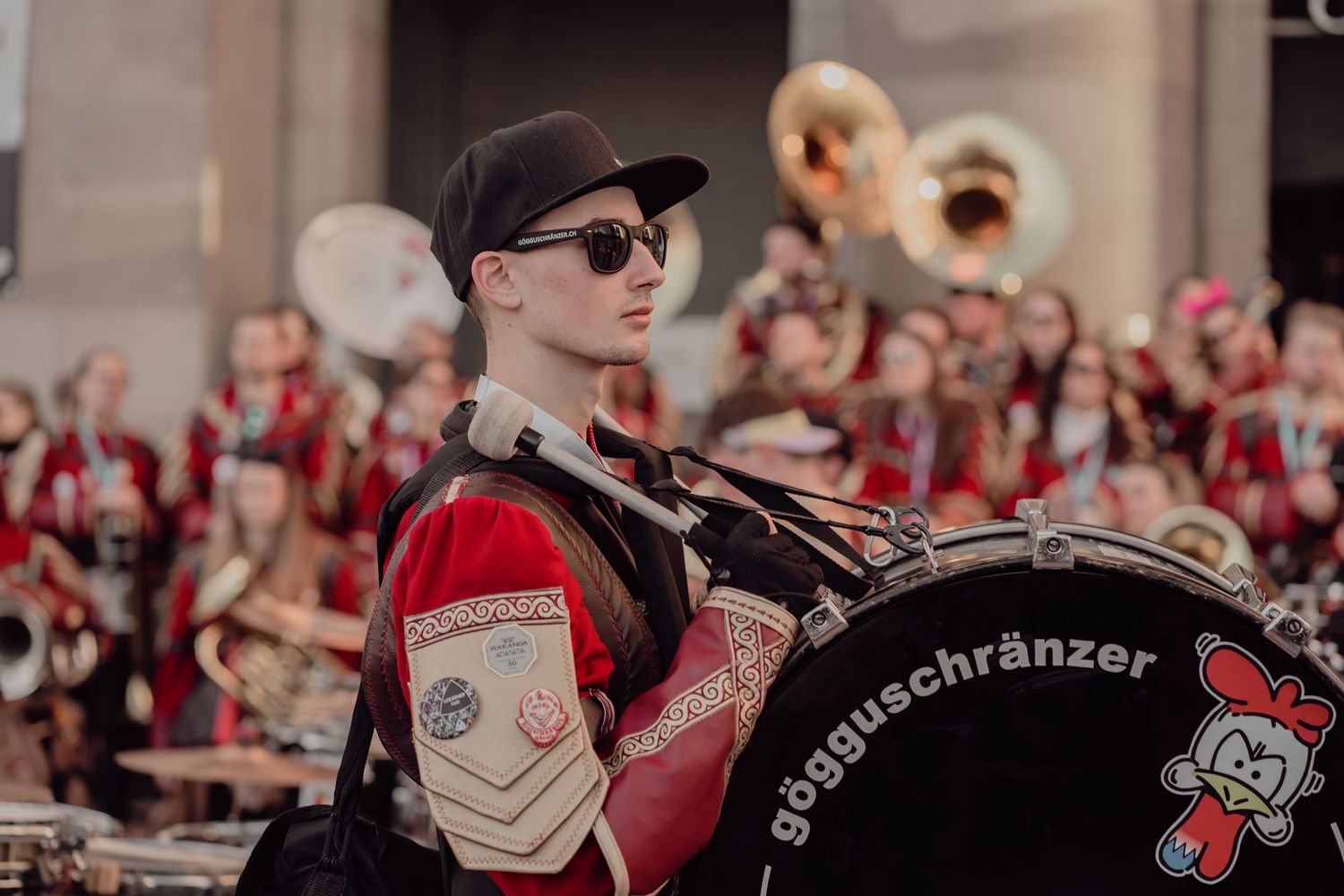 A young drummer in red and black uniform with sunglasses plays bass drum at a parade.