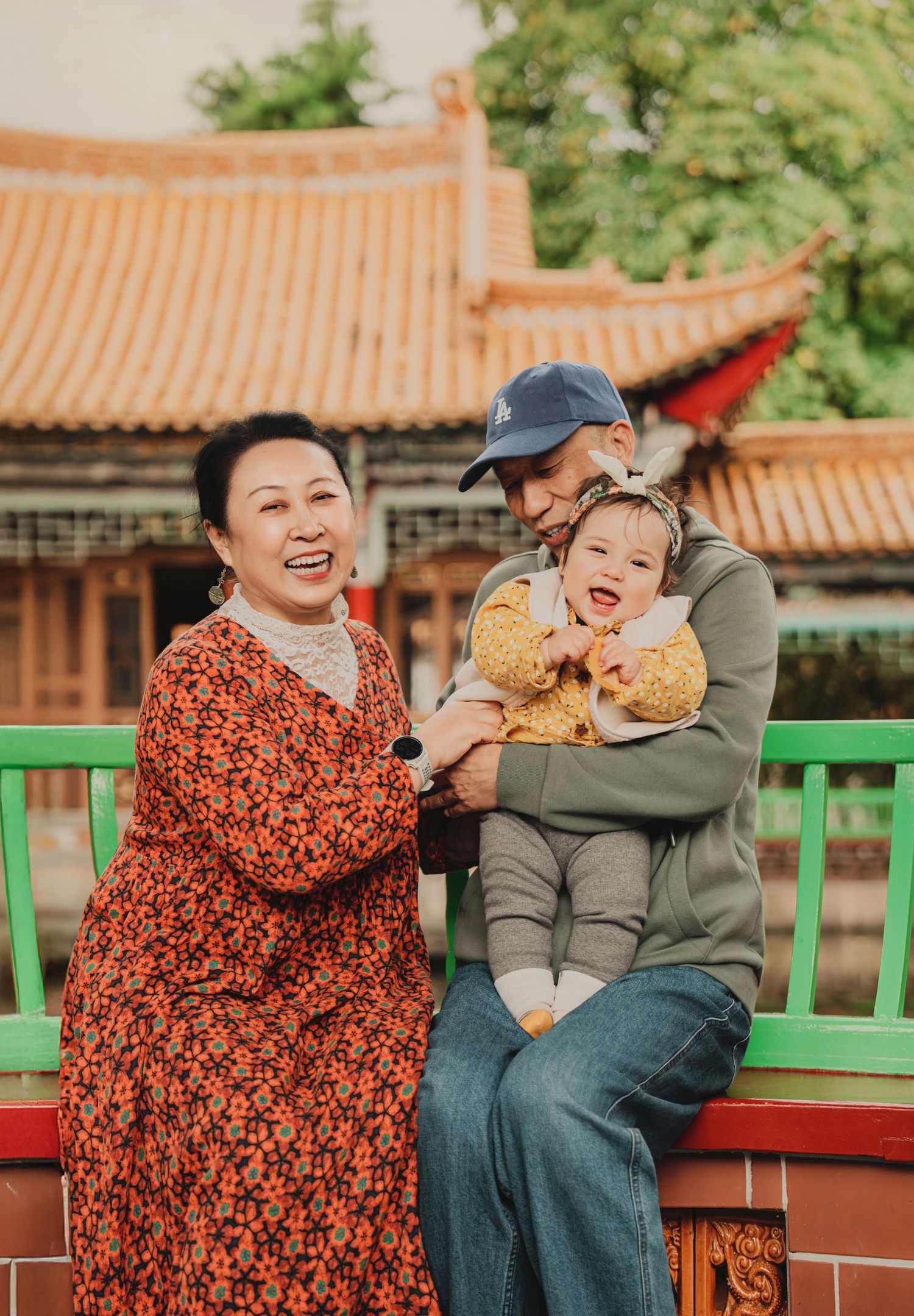Grandparents laughing with a baby girl in yellow at a colorful Chinese garden pavilion.