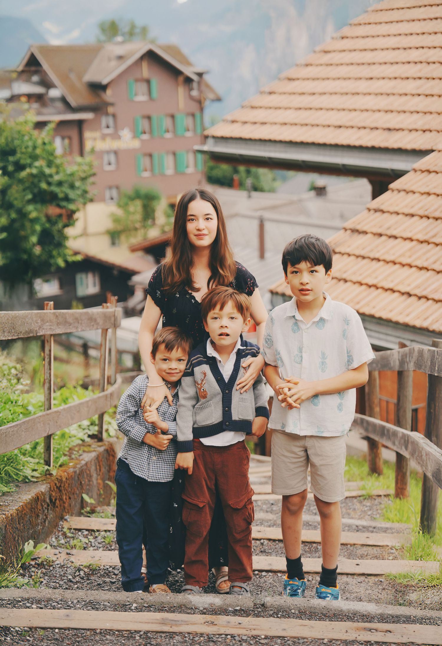 Asian mother poses with three sons on wooden stairs in a scenic Swiss alpine village.