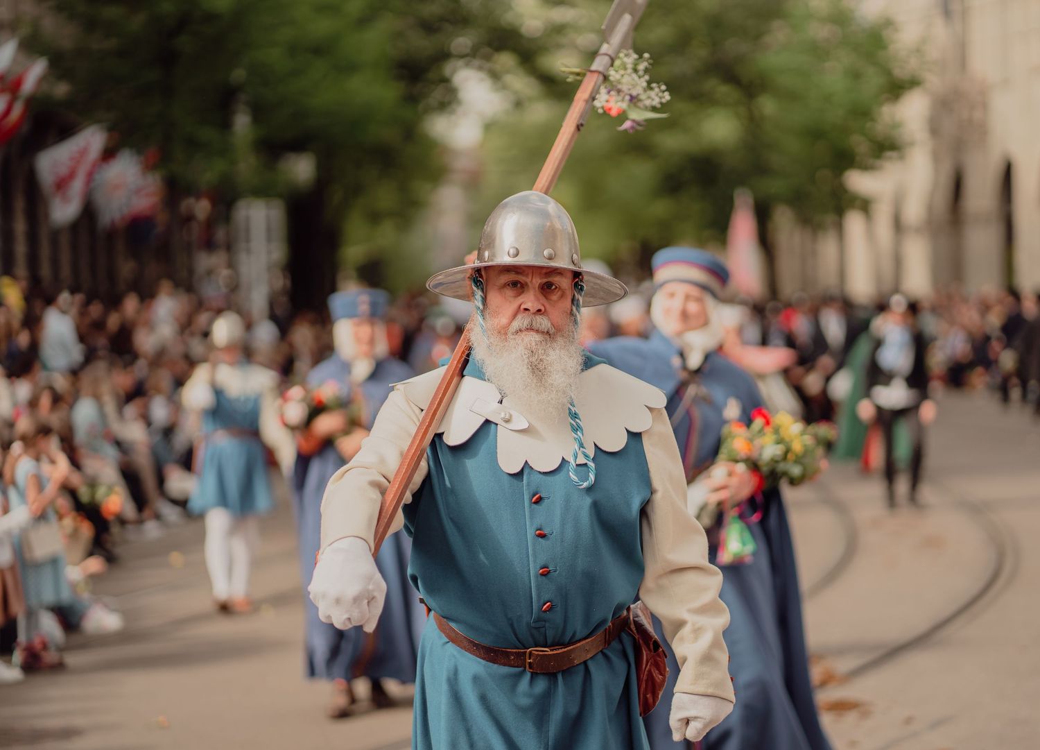 An elderly man in a blue historical costume with a metal helmet carries a staff in a city parade.