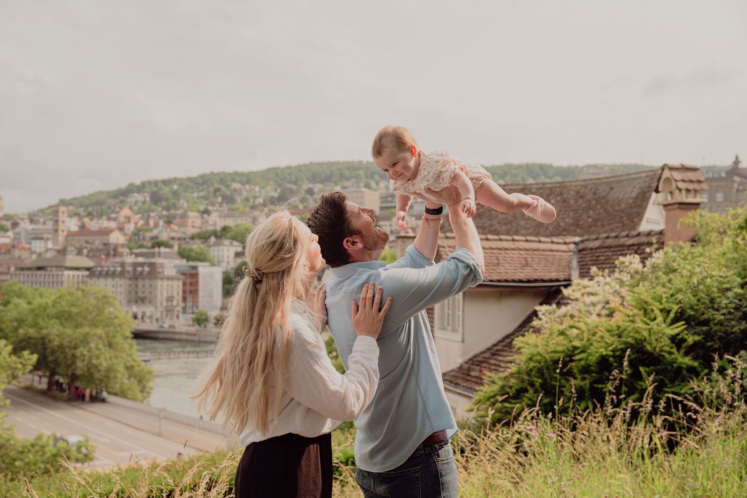 Family of three outdoors, father lifting baby in air while mother embraces him, European cityscape background.