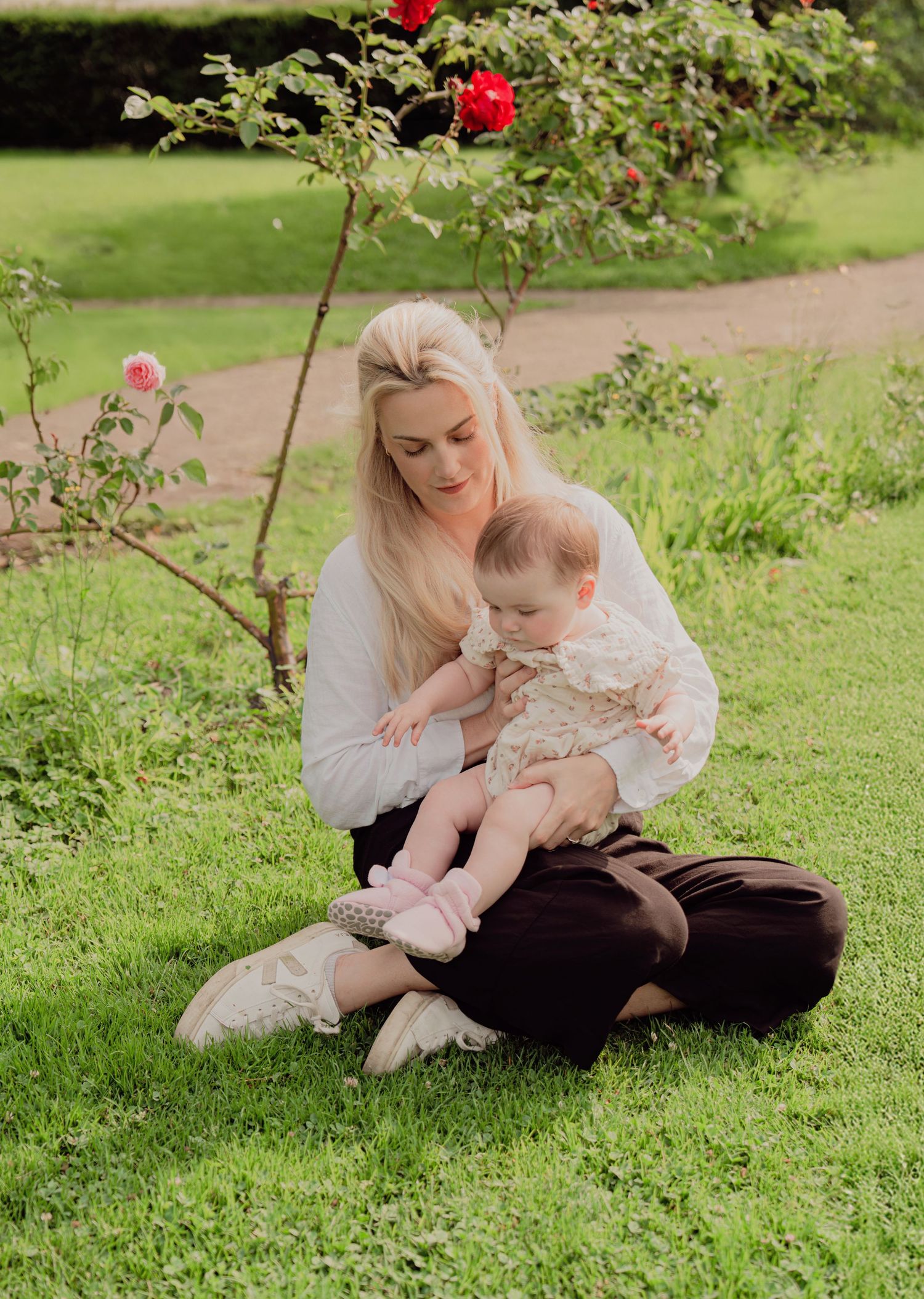Blonde mother sitting cross-legged on grass cradling baby in floral outfit near rose bush.
