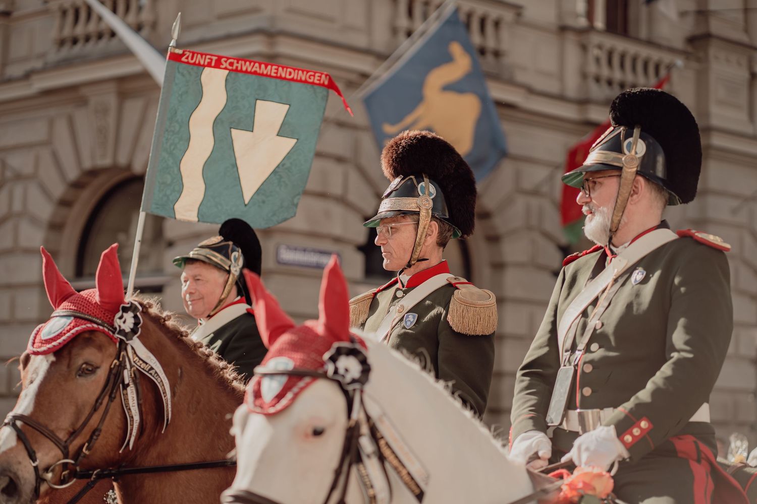 Three riders in historical military uniforms on horseback carry a banner at a traditional city parade.